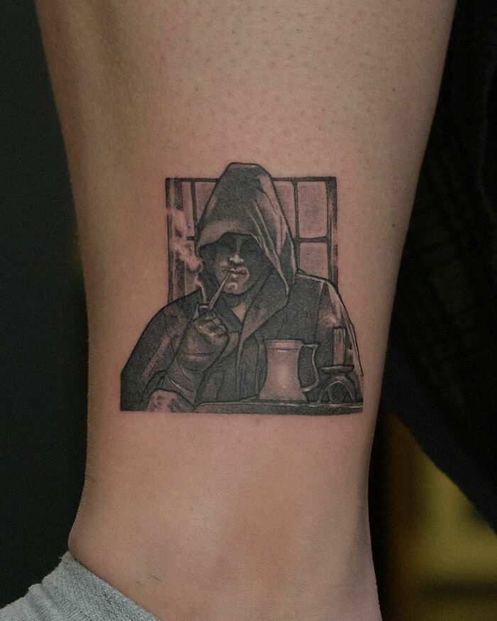 Hooded figure from Lord of the Rings tattooed on leg, smoking a pipe in a shaded design.