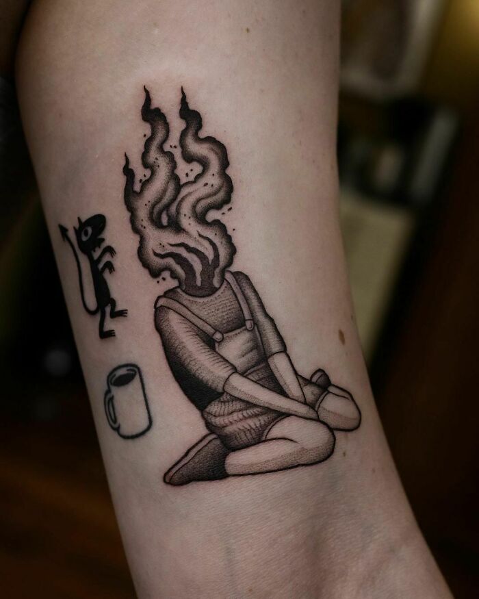 Tattoo of a person with a fiery head, kneeling beside a cat and a mug, symbolizing mental health healing.