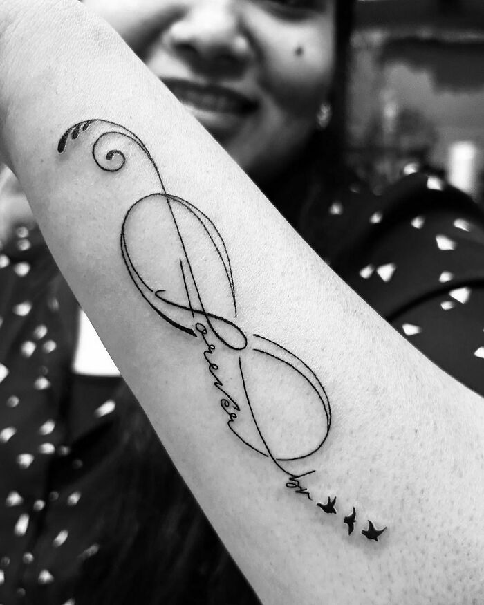 Infinity tattoo with 'forever by' text and birds, symbolizing eternal creativity on a person's arm.