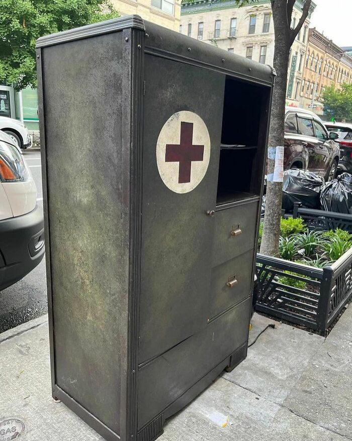 Old medical cabinet with a red cross symbol discarded on a NYC sidewalk among other trash and parked cars.