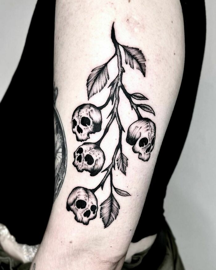 Gothic tattoo of skulls and leaves on an arm, showcasing intricate design and dark elegance.