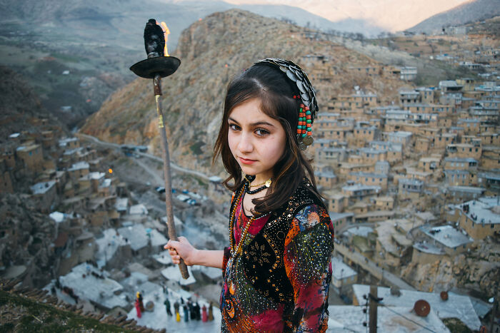 Young girl in traditional clothing holds a torch with a mountainous village background in best pictures chosen by community.
