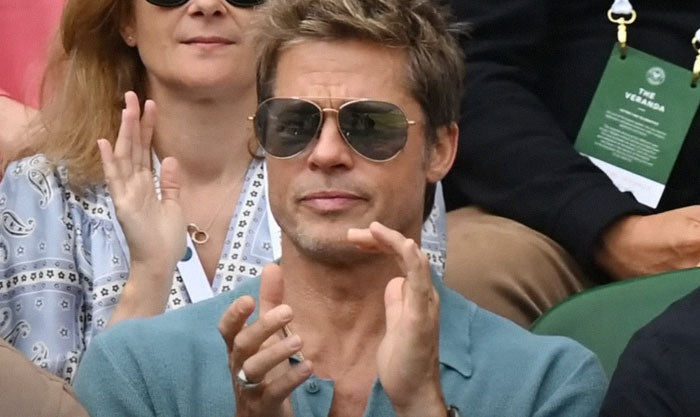 Brad Pitt at Wimbledon, wearing sunglasses and a teal shirt, clapping during the match.