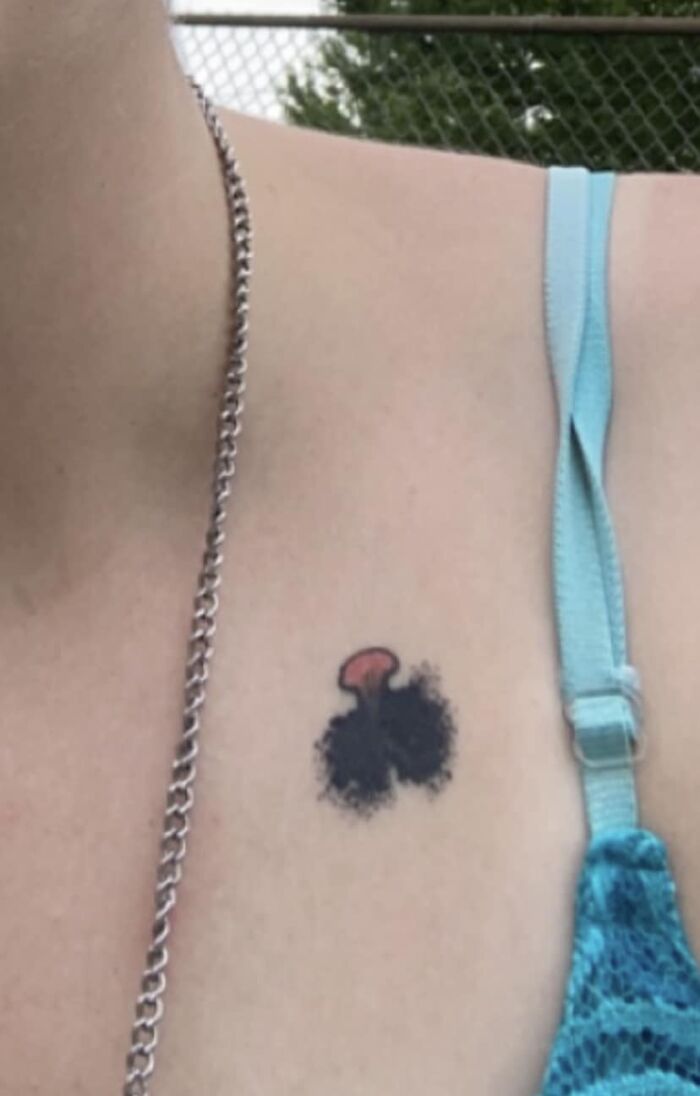 Funny tattoo of a black cloud with a small red shape on a person’s upper chest, near a blue strap and silver chain.