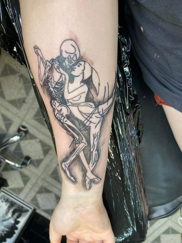 Tattoo of a skeleton and a woman kissing on a person's forearm, showcasing a humorous or unusual design.