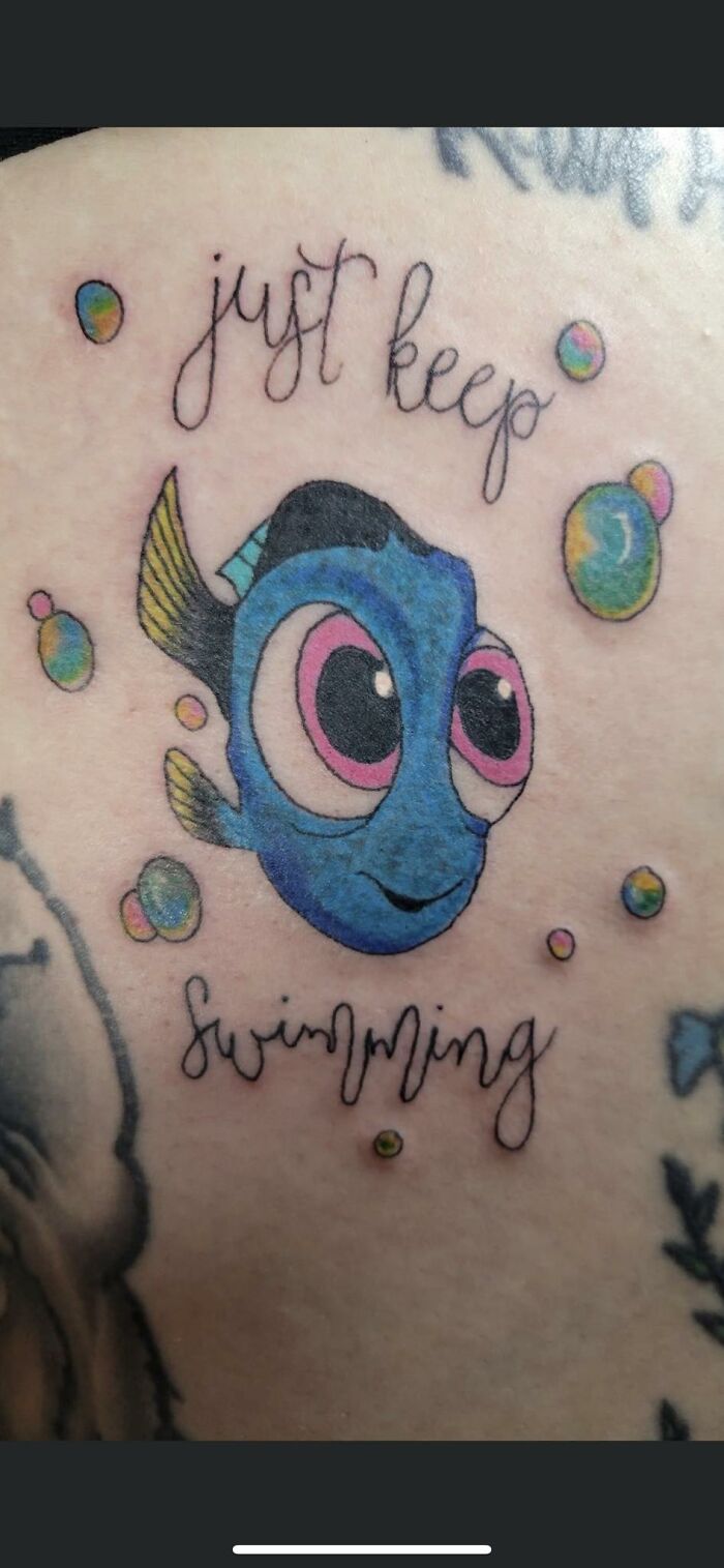 Funny tattoo of a blue fish with big eyes and the phrase "just keep swimming" surrounded by colorful bubbles.