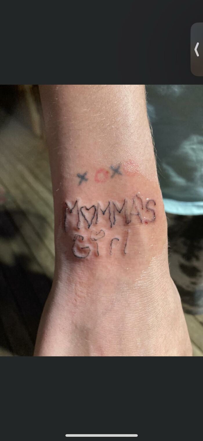 Funny tattoo reading "Momma's Girl" on a wrist with uneven handwriting and hearts around it.
