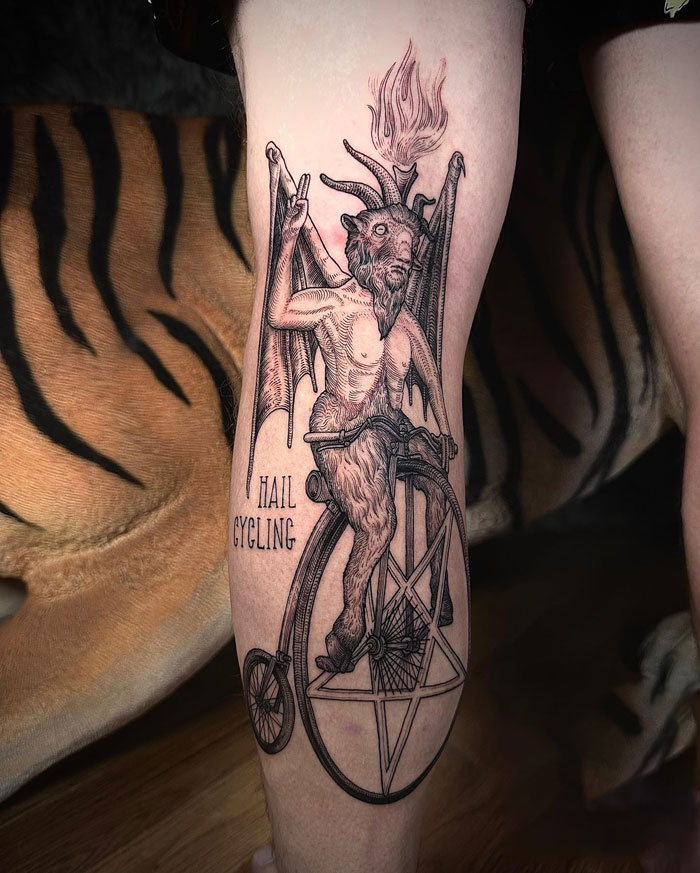 Gothic tattoo of a satyr on a bicycle with a pentagram, highlighted by intricate details and bold design on a person's leg.