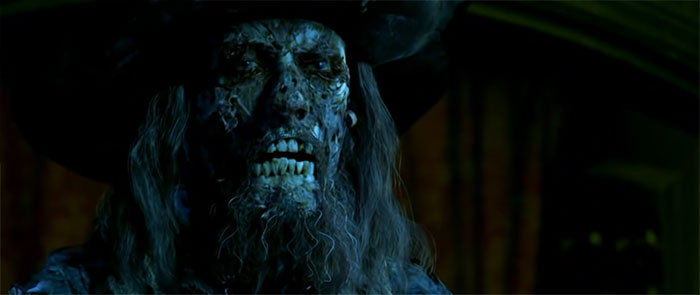 Hector Barbossa with decayed face and pirate hat staring intensely in a dark scene from Pirates of the Caribbean quotes.