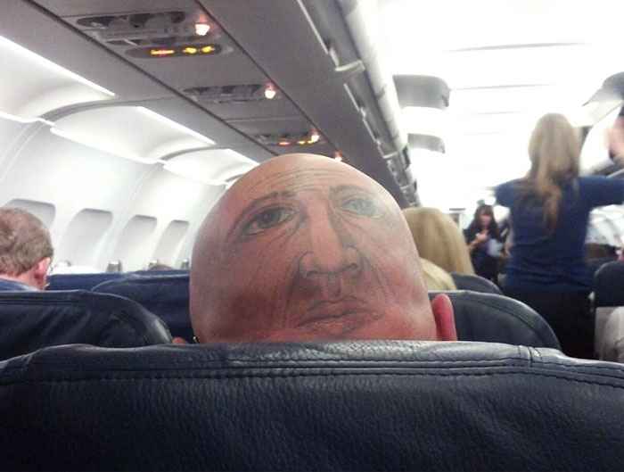 Unusual tattoo of a face on a bald head seen inside an airplane cabin.