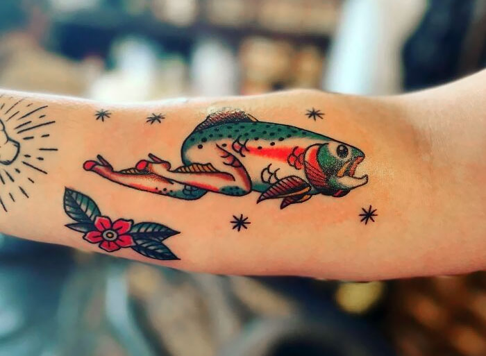 Colorful tattoo of a fish combined with a human leg on an arm, accompanied by small stars and a flower.