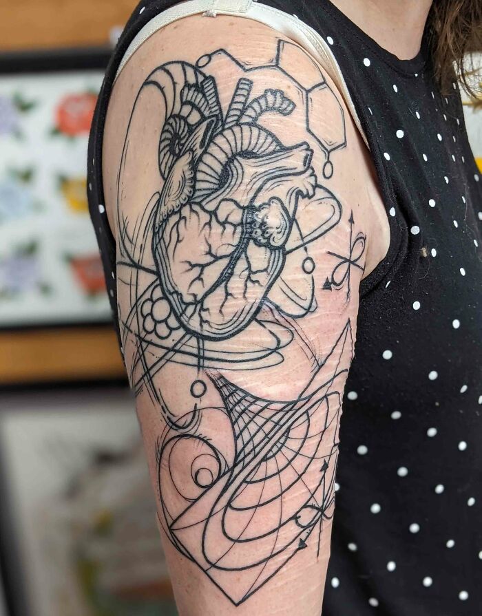 Science tattoo featuring a heart, atomic symbols, and geometric patterns on a person's upper arm.