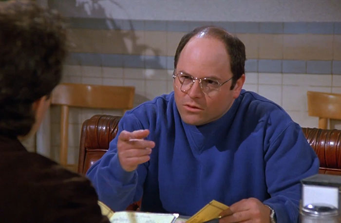 George wearing a blue sweater, pointing and talking during a scene featuring funniest Seinfeld quotes about everyday life.