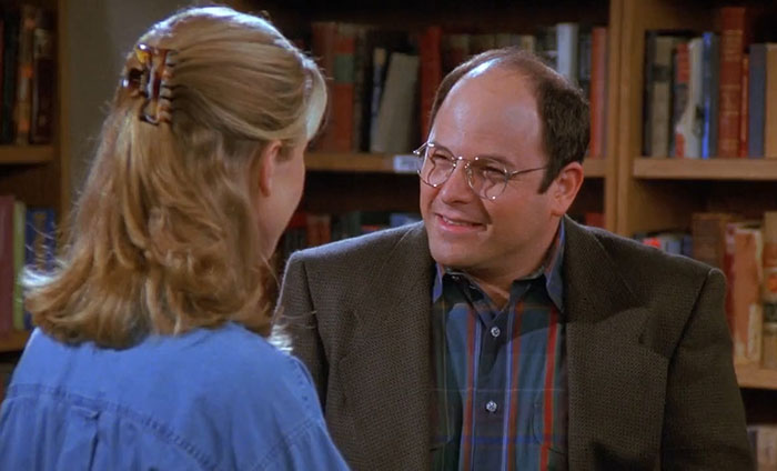 George wearing a brown suit and colorful shirt in a library setting, illustrating funniest Seinfeld quotes about everyday life.