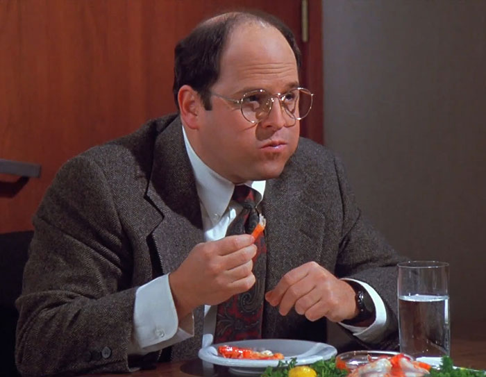 George wearing a gray brown suit eating shrimp at a table, capturing funniest Seinfeld quotes about everyday life moments.