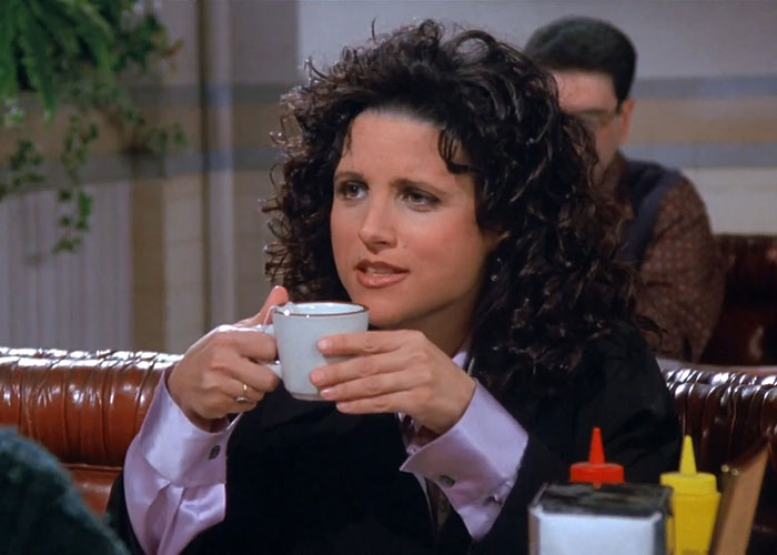 Elaine from Seinfeld wearing black suit and purple shirt, holding a coffee cup in a diner scene about funniest Seinfeld quotes.
