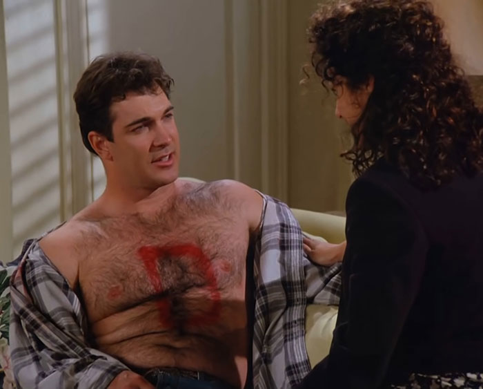 David Puddy from Seinfeld wearing a plaid shirt opened, showing a painted chest emblem in a humorous everyday life moment.