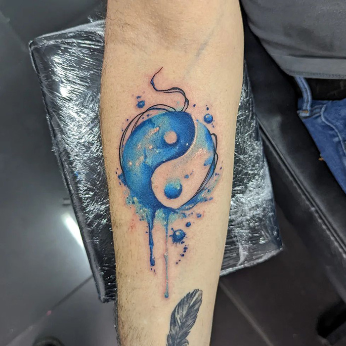 Blue Yin Yang tattoo with watercolor effect on forearm, symbolizing life's duality.