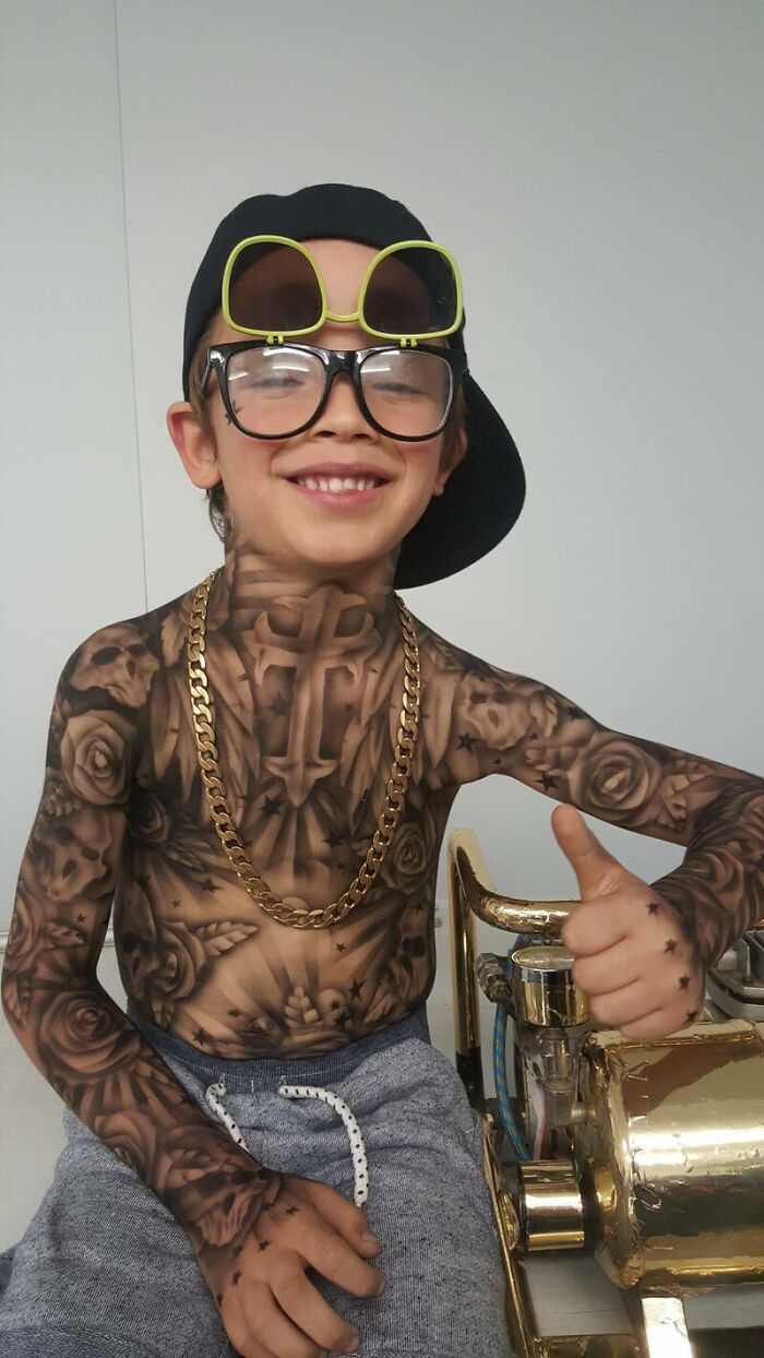 Child with spray-on tattoos, wearing oversized glasses and a black cap, smiling and giving a thumbs up.