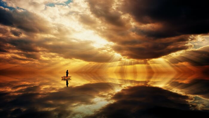 Person standing alone on a small boat surrounded by reflective water under dramatic deep sky and glowing light rays.