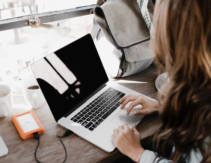 Woman using laptop at a wooden table with external hard drive and backpack near window, symbolizing grandchild inheritance issues.