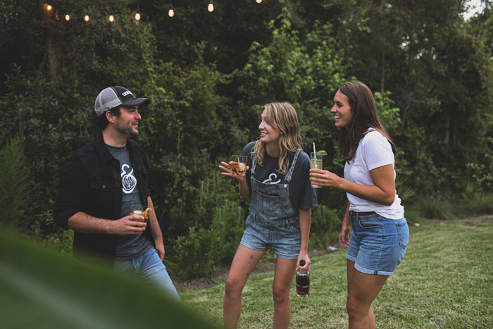 Three friends laughing outdoors, holding drinks, with trees and string lights in the background. Three friends laughing outdoors, holding drinks, with trees and string lights in the background.
