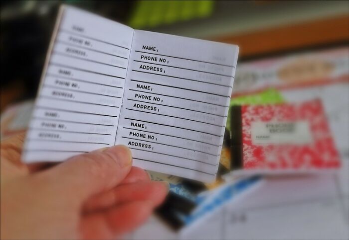 Hand holding a small blank address book with columns for names, phone numbers, and addresses in focus.