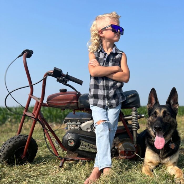 Child with Cheddar Wiz mullet hairstyle wearing sunglasses, standing by a minibike with a dog on grassy field under clear sky.