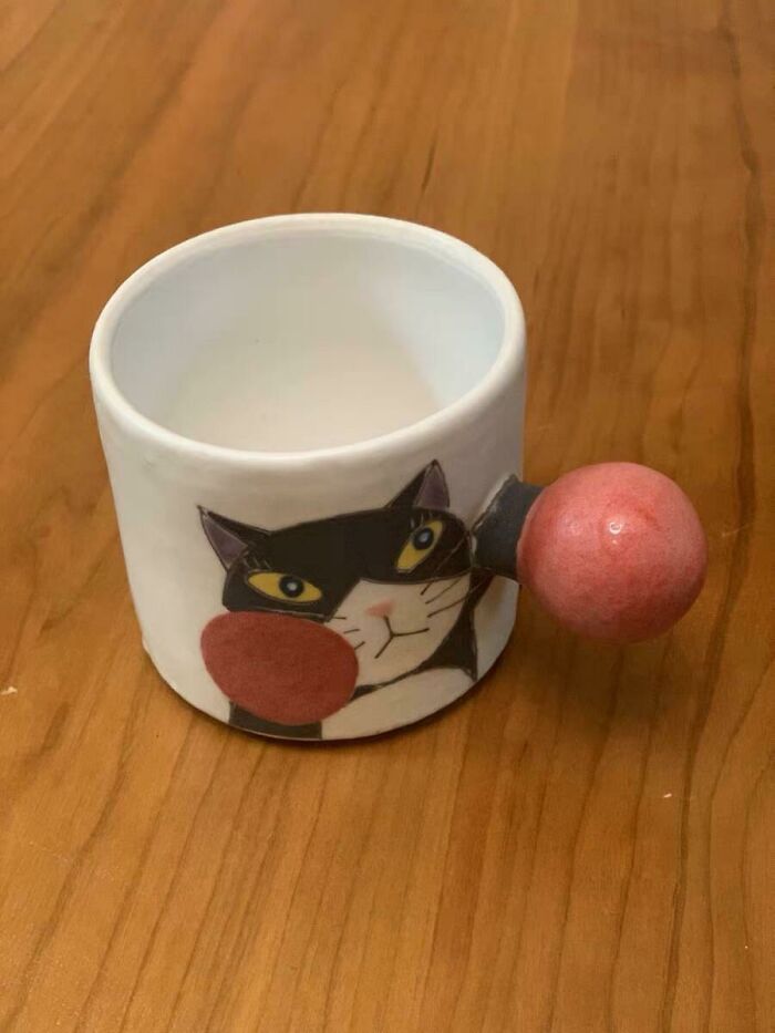 Ceramic mug with a cat face design and a unique round handle resting on a wooden surface, showcasing mug life creativity.