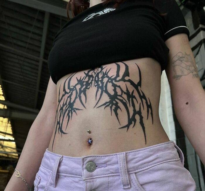 Woman with a tribal tattoo across her stomach, wearing a black top and pink pants, showcasing body art inspiration.