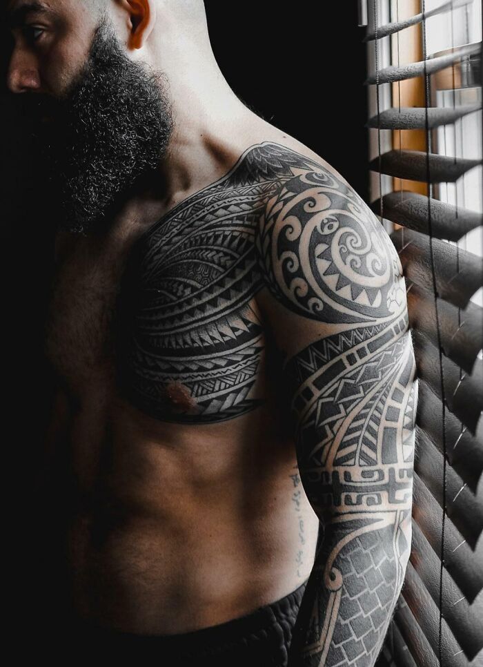 Man with intricate tribal tattoos on shoulder and arm, standing in soft light near window blinds.