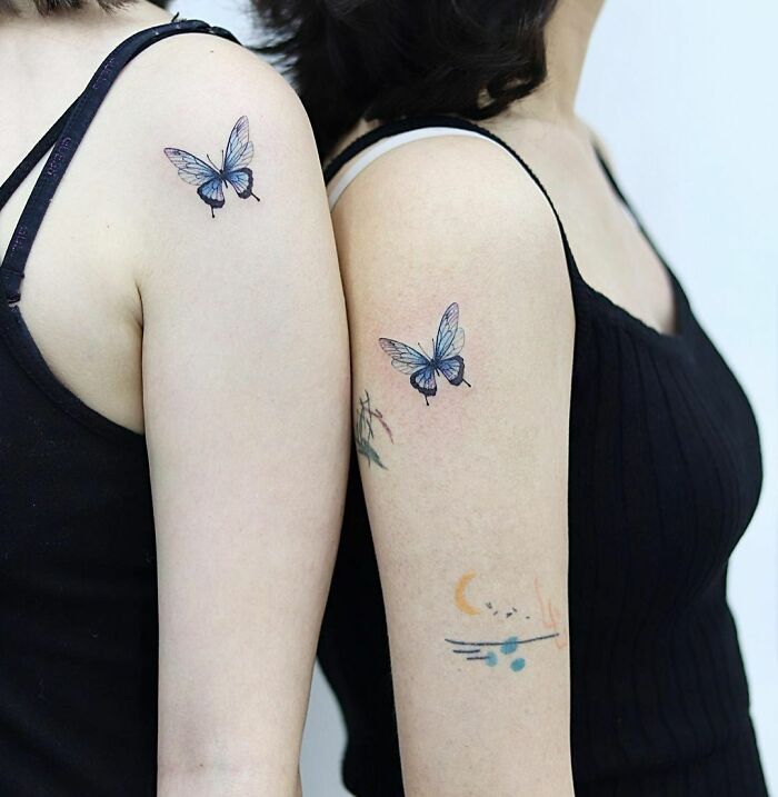 Two women showing off colorful butterfly tattoos on their upper arms.