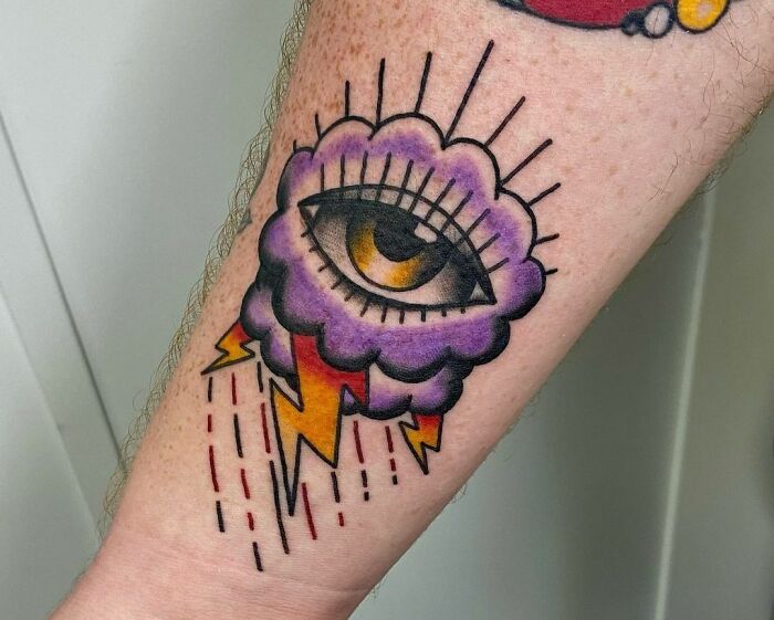 American traditional tattoo of an eye in a purple storm cloud with yellow lightning bolts and red rain on a forearm.