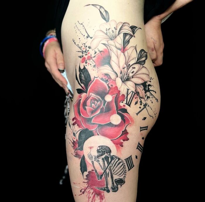 Trash Polka tattoo on thigh, featuring roses, lilies, a skeleton, and clock elements in red and black.