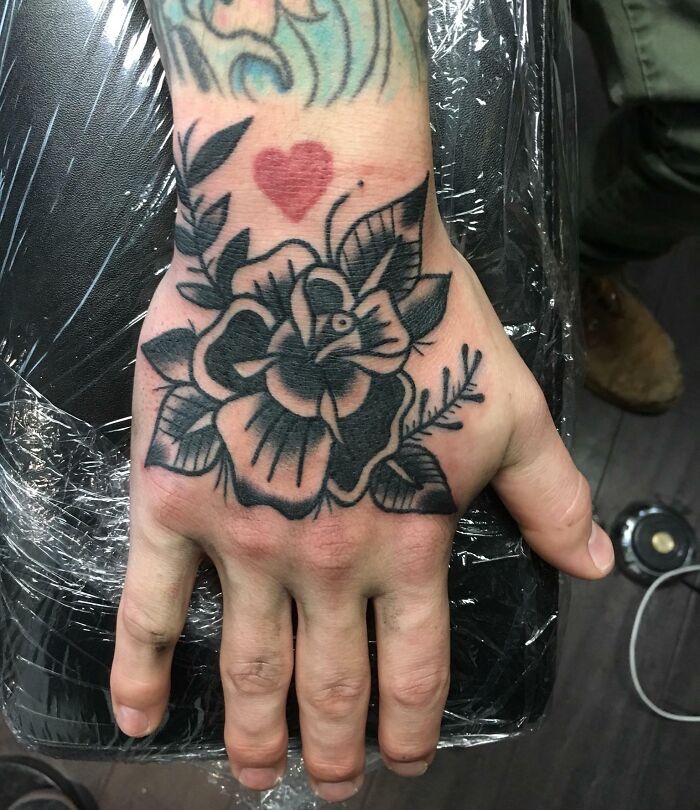 Hand displaying an American traditional tattoo of a rose and heart, showcasing bold black outlines and classic design elements.