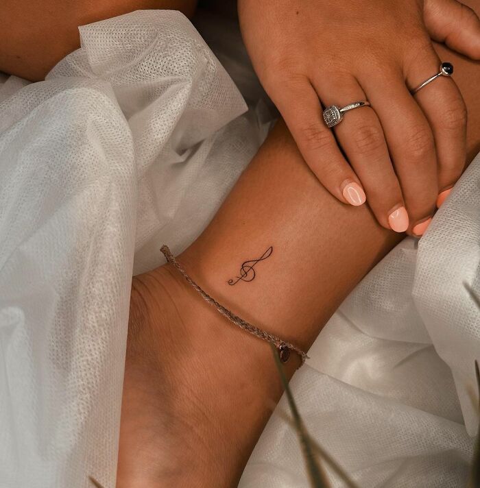 Ankle tattoo of a treble clef with a chain bracelet, surrounded by white fabric, illustrating ankle tattoo care.