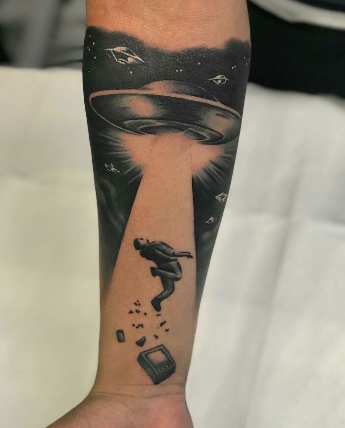 Tattoo of a UFO beaming up a person, with space elements, on a forearm.