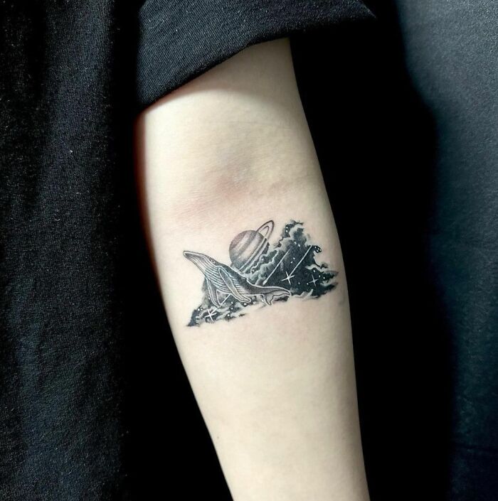 A cool space tattoo featuring a whale, Saturn, and stars on a forearm.