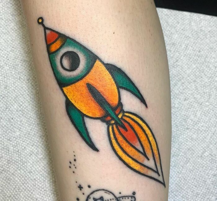 Colorful rocket space tattoo on arm, featuring orange, yellow, and green hues with flame-like detail.