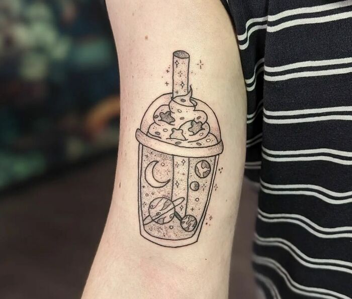 Space tattoo of a boba drink with planets and stars, on a person's arm wearing a striped shirt.