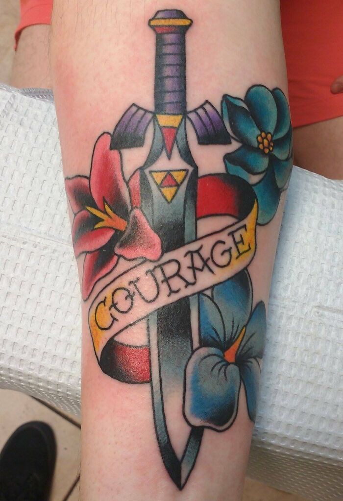American traditional tattoo with a sword, flowers, and a "Courage" banner, showcasing bold colors and classic design.