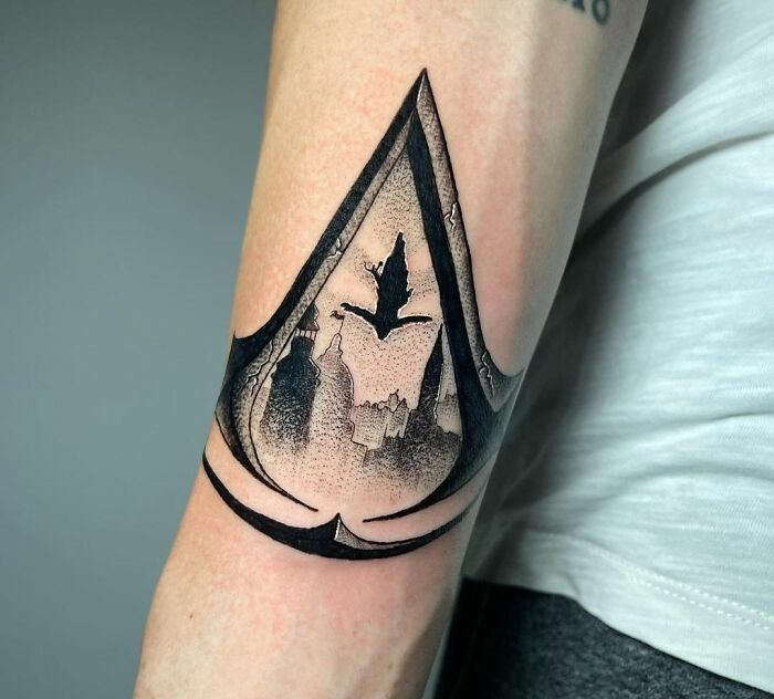 Assassin's Creed video game tattoo with a stylized symbol, featuring a cityscape and a flying bird design on a forearm.