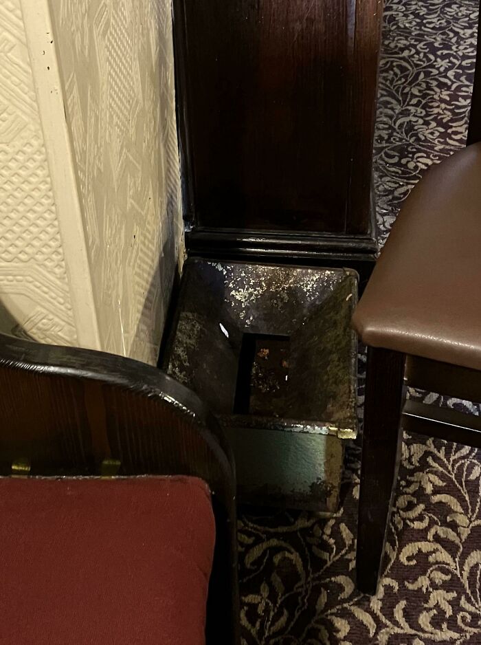 Rusty metal object with a square hole, placed between chairs on a patterned carpet, a mysterious object found indoors.
