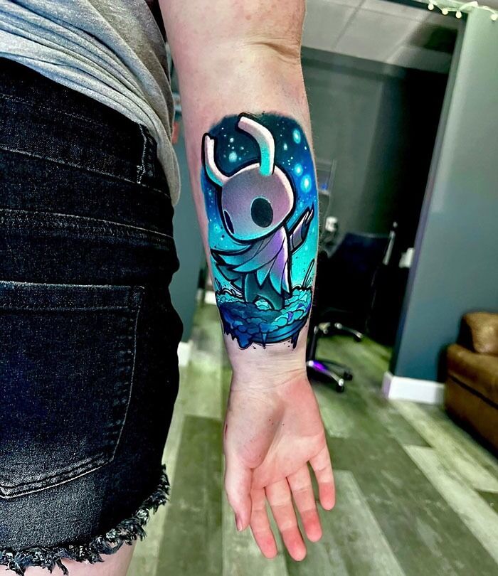 Gamer tattoo of a Hollow Knight character from a video game on a person's forearm, vibrant colors highlighting the design.