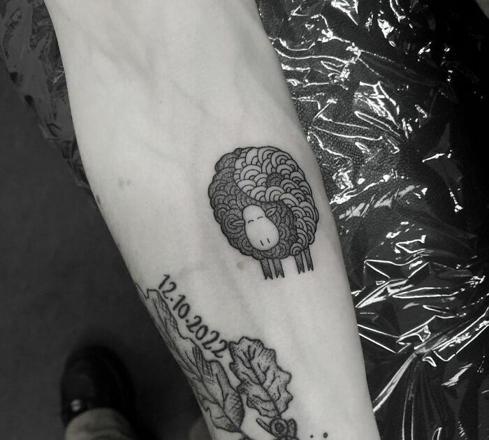 Black and gray sheep tattoo with date and leaves, symbolizing yin yang duality on forearm.