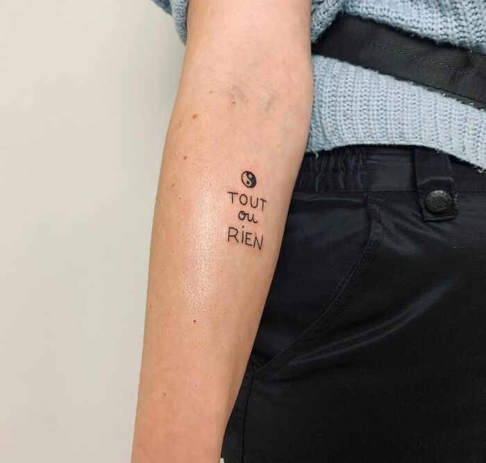 Yin Yang tattoo on arm with "Tout ou Rien" text, symbolizing duality.