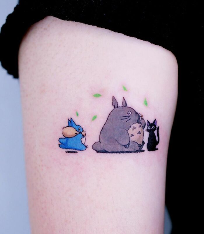 Cartoon tattoo featuring animated characters from My Neighbor Totoro with a blue creature, a gray creature, and a small black cat on skin.