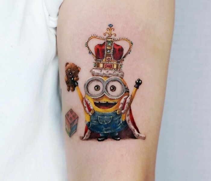 Cartoon tattoo of a crowned minion wearing a robe, holding a teddy bear, inspired by animated shows.