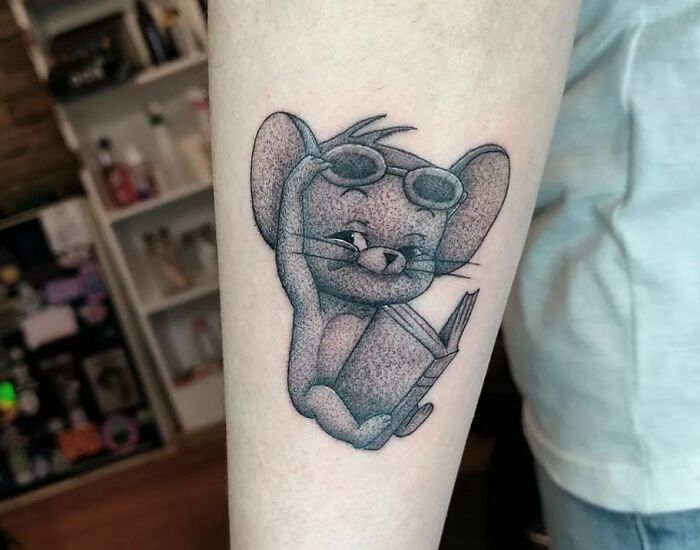 Cartoon tattoo of a mouse with sunglasses, holding a book, inked on forearm, inspired by animated shows.