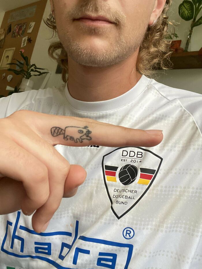 Person pointing at a small, comically drawn tattoo on their finger, highlighting the choice of tattoo artist.